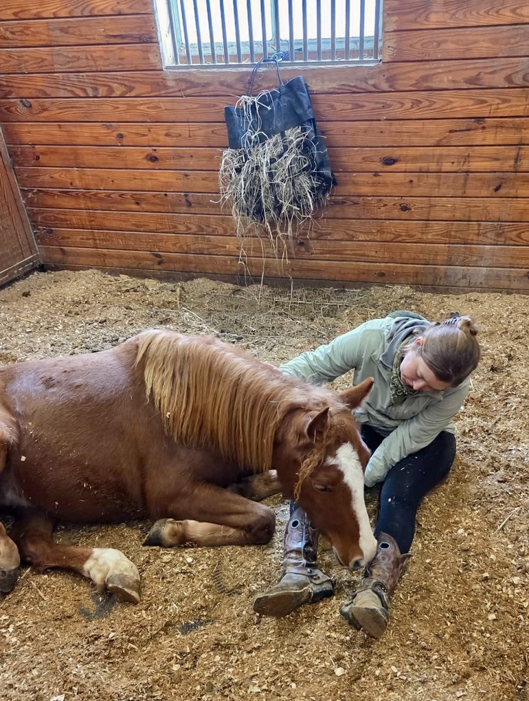 Meg sitting in the stall bedding beside a resting chestnut horse, its head against her boots