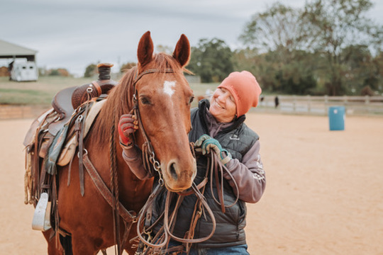 A handler in a beanie sharing a moment with a saddled chestnut horse